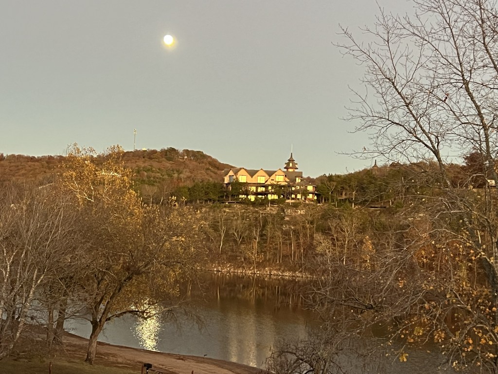 A scenic view of a riverbank with bare trees in the foreground, illuminated by the glow of a full moon in the sky. In the background, a large house with multiple gables sits atop a hill surrounded by tree-covered land.