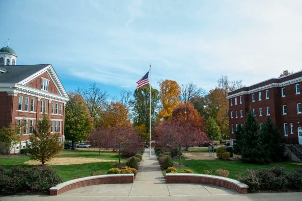 A college campus scene featuring a pathway leading to a flagpole with an American flag, surrounded by autumn foliage and brick buildings.