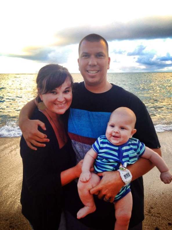 A happy family portrait by the beach, featuring a woman and a man embracing each other while holding a smiling baby. The backdrop shows water and a colorful sky.