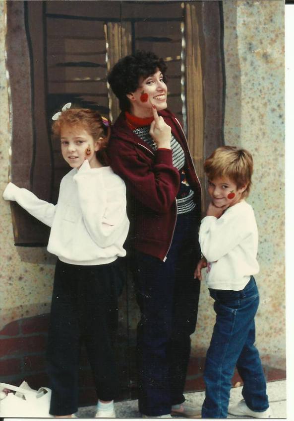A playful photo of a woman and two children, all making silly faces. The woman is in a dark jacket, while the children are in white tops, with the girl on the left having red face paint. They are posing in front of a backdrop resembling a house with a window.