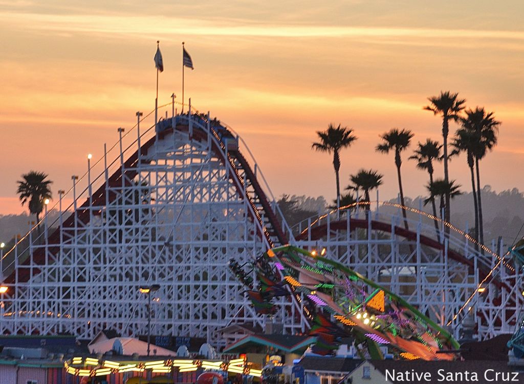 A sunset view of a wooden roller coaster with palm trees in the background, located at an amusement park.